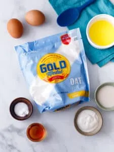 table with bag of oat flour, two eggs, pot of honey, bowl of plant butter, and cup of plant milk with a mixing spoon