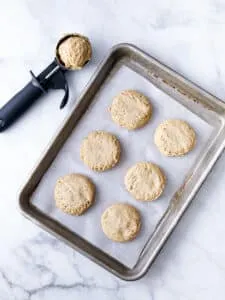 baking sheet covered in parchment paper with 6 drop biscuits padded out with a cookie scoop next to the tray