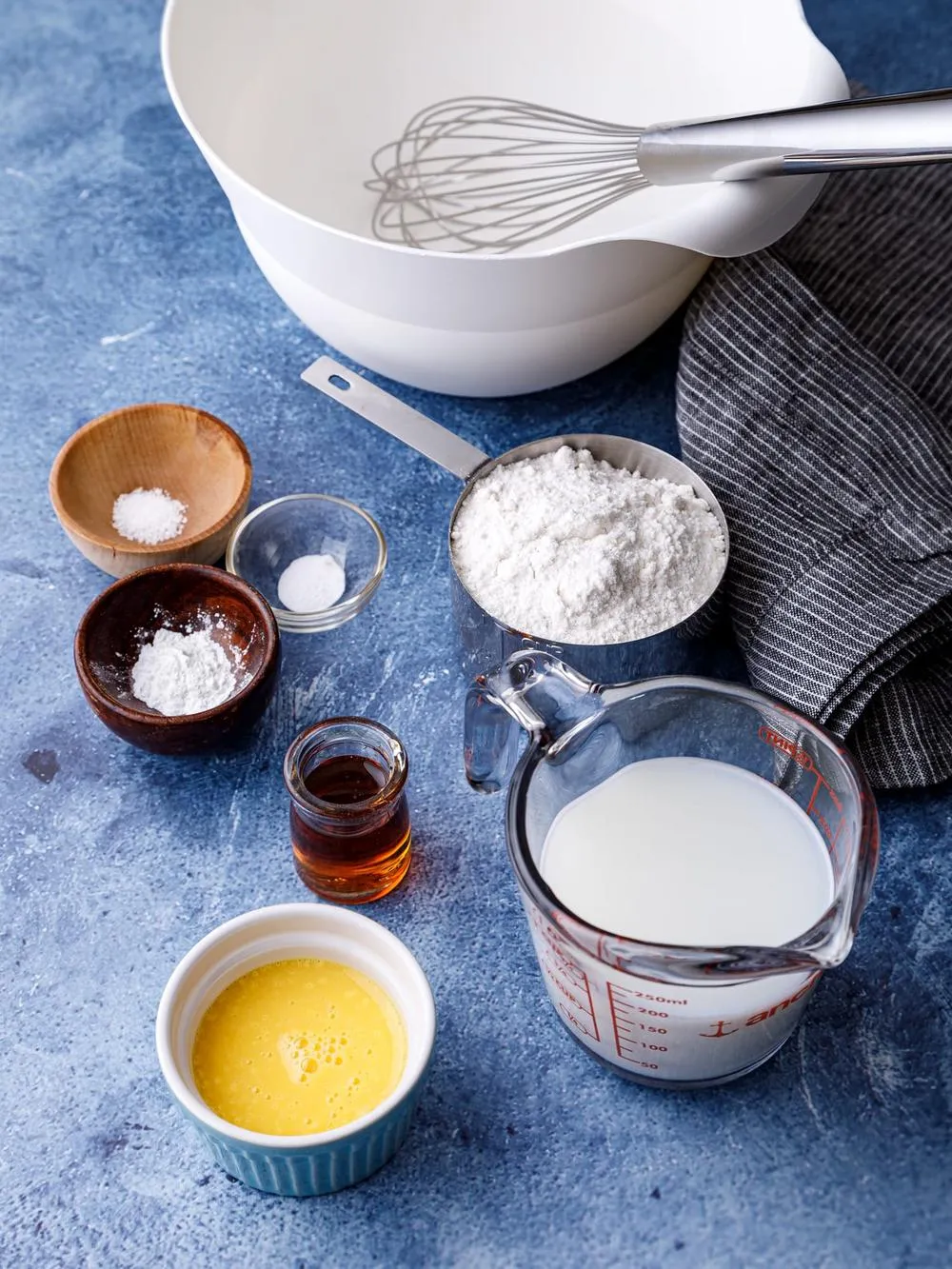 White bowl with a whisk a measuring cup of gluten free flour and almond milk a bowl of egg substitute, vanilla and baking soda on a table with a dishcloth