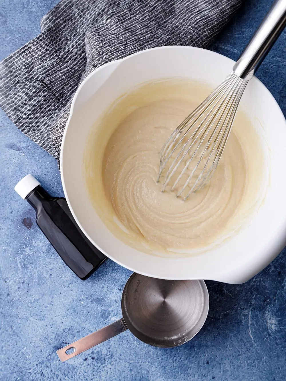 Mixing bowl of almond milk pancake batter with a silver whisk on a table with a measuring cup and bottle of vanilla on a table with a dishcloth