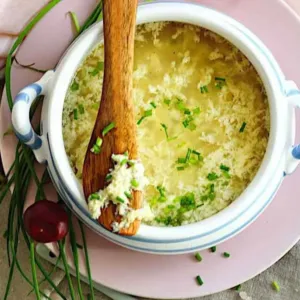 white bowl of Keto Egg Drop Soup on a pink plate with a wooden spoon and herbs