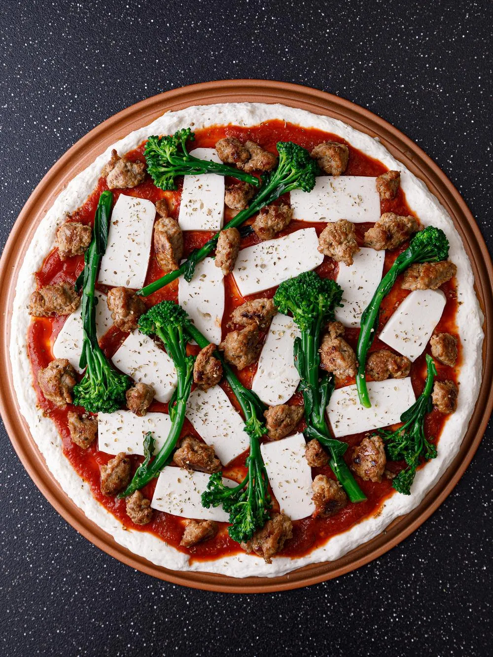 a gluten free pizza crust topped with no tomato pizza sauce, broccolini, ground turkey and vegan mozzarella on a black tabletop